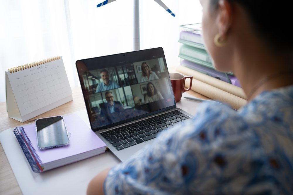 Woman in a blue patterned shirt participates in a video call at her home office desk with a laptop, notebooks, calendar, and coffee mug, telecommuting, Spain.
