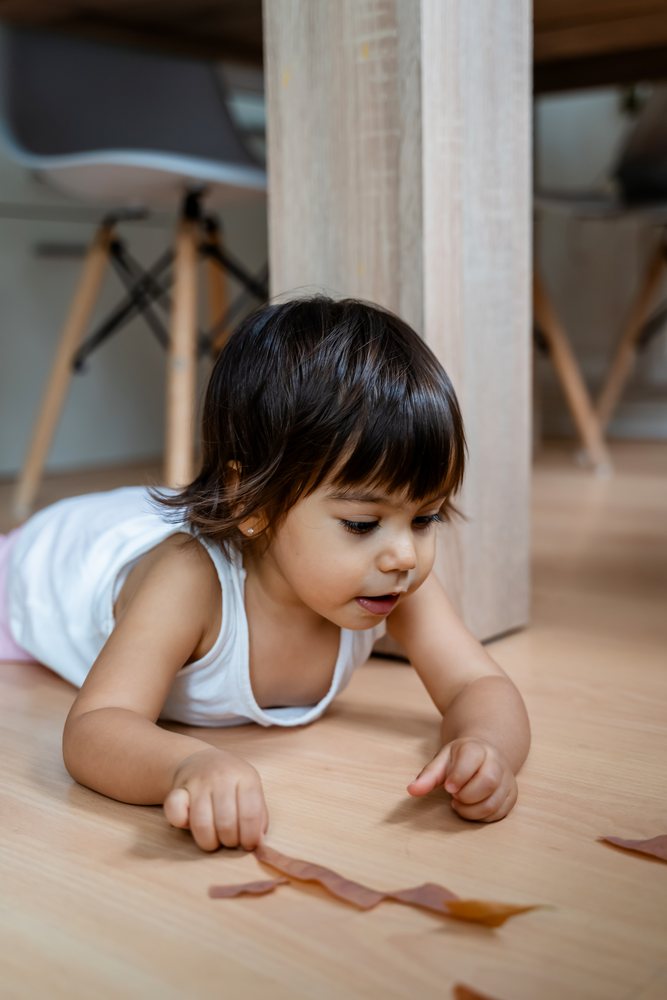 Toddler playing with pieces of paper on a wooden floor under a light wood table indoors, kids, Spain.