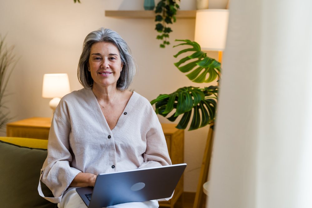 Middle-aged woman with gray hair sits and smiles while using a laptop in a living room with indoor plants and lamps, Spain.