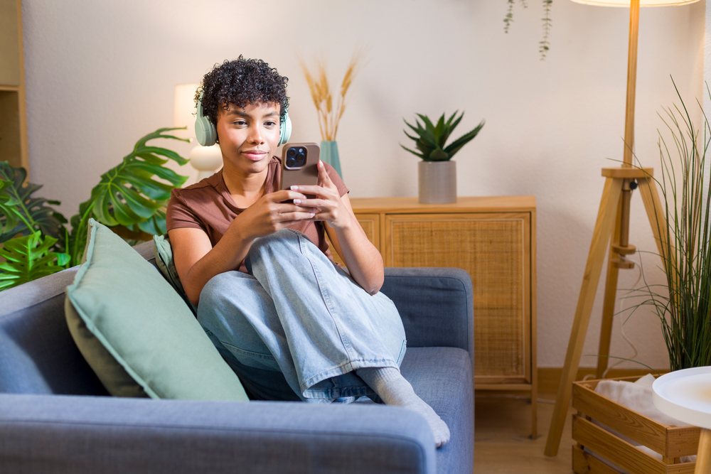 Young woman with curly hair uses a smartphone while sitting cross-legged on a sofa in a modern, cozy living room.