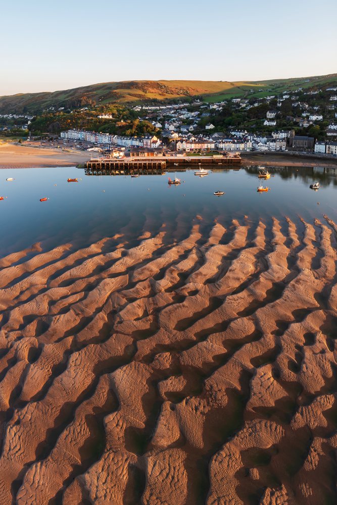 Sand and Mud form ripple patterns at the Dyfi Estuary in Ynyslas, with boats, a pier, and a coastal town visible under sunlight reflection, Landscape, Aberdyfi, Gwynedd, Wales.