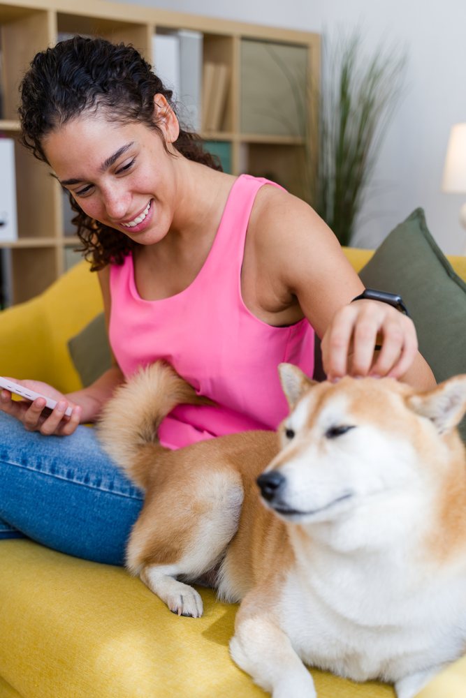 A woman in a pink tank top smiles while petting a dog sitting on a yellow couch in a living room setting, Spain.