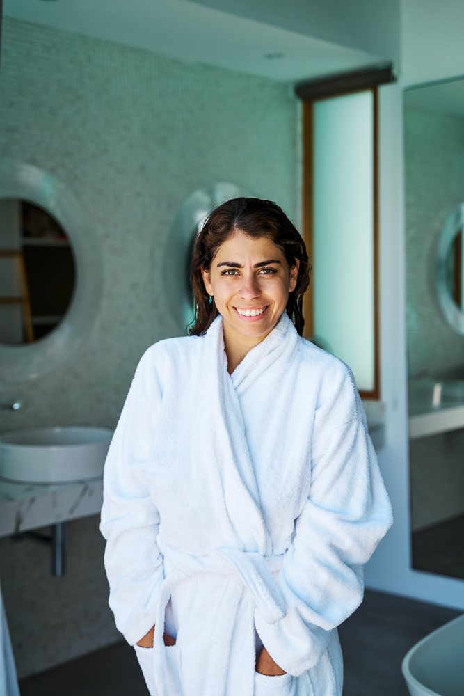 A smiling woman wearing a white bathrobe stands in a modern bathroom with circular mirrors and marble countertops in the, Maldives.