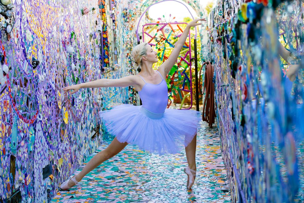 Female ballet dancer in a lavender tutu poses expressively amid colorful mosaic walls and floor in an outdoor artistic space, CA, USA.