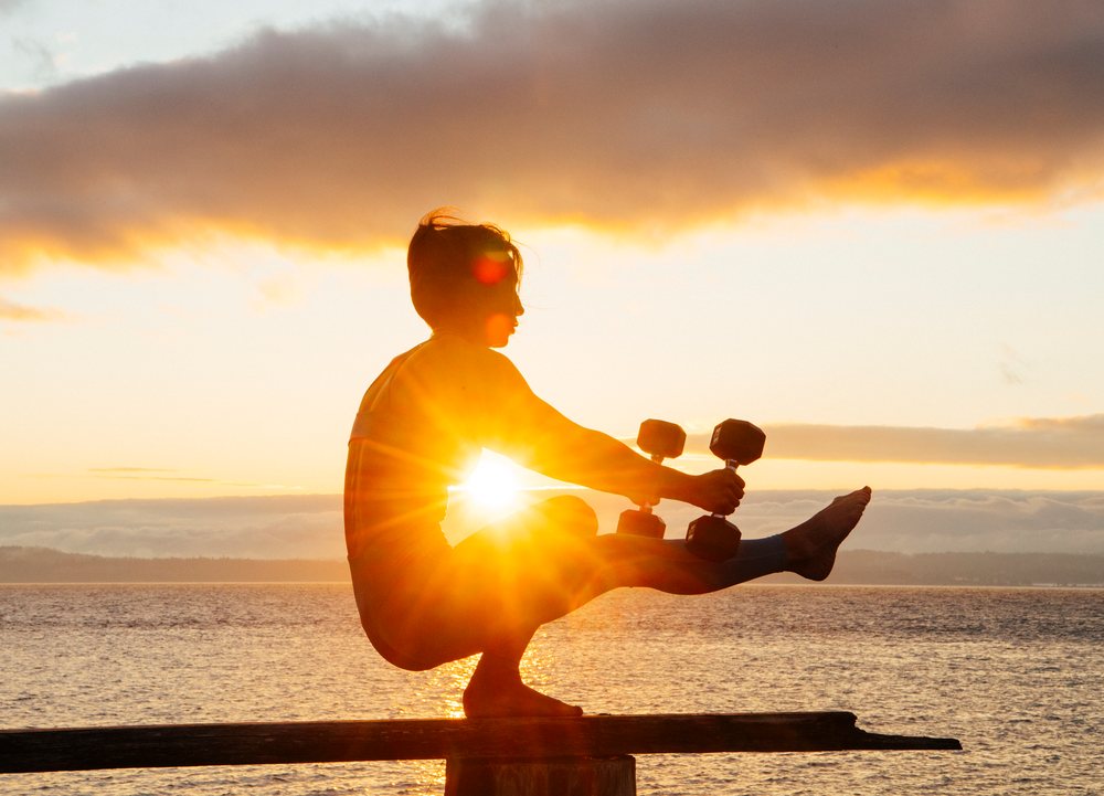 Silhouette of a barefoot woman balancing on a wooden beam holding dumbbells at sunset by the water, WA, USA.