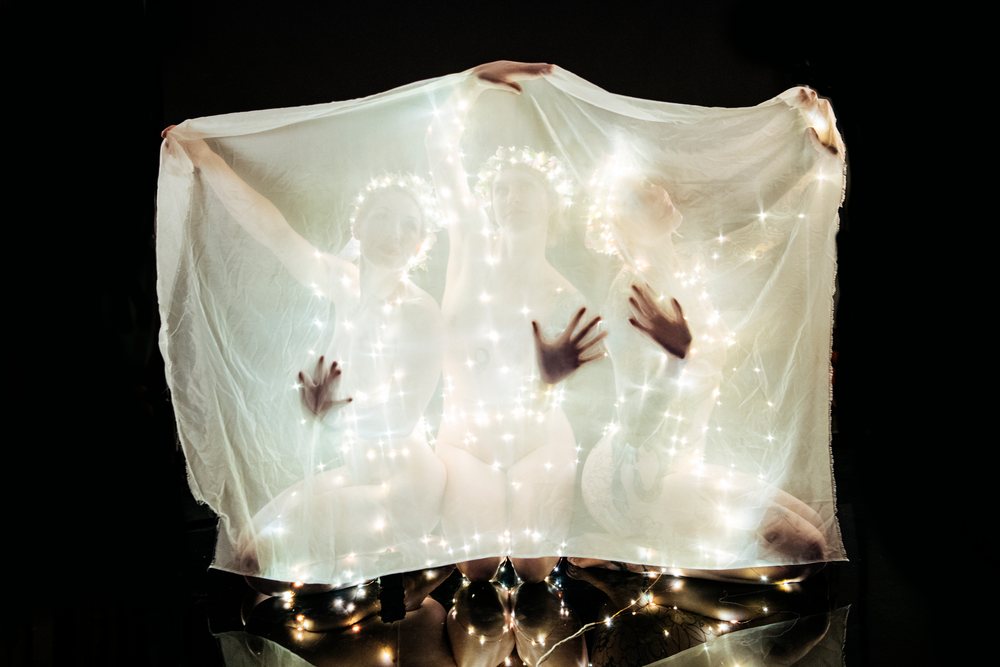 Three kneeling women hold a translucent veil decorated with glowing fairy lights and wear flower crowns against a black background, muses, USA.