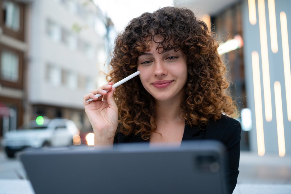 Smiling woman with curly hair uses a digital tablet and stylus in an urban Madrid setting, young business woman.