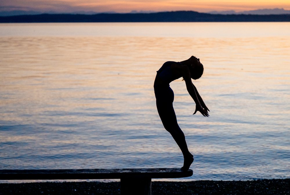 Woman practicing yoga acrobatics with a deep backbend silhouette by a lake at sunset, USA.