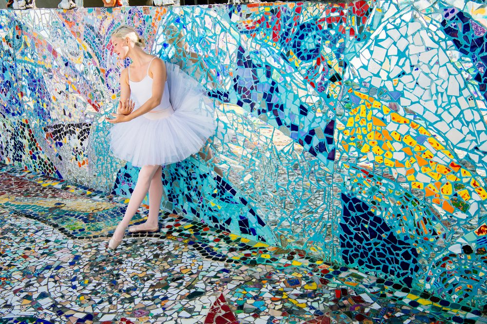 Ballerina in white tutu and ballet shoes performing beside a colorful mosaic wall reflecting her image in an urban outdoor art setting, CA, USA.