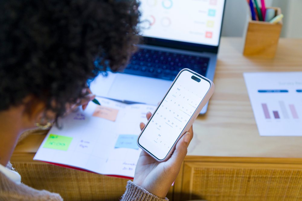 Woman in a home office planning her schedule using a smartphone calendar app, laptop, and notebook with pen, remote work.