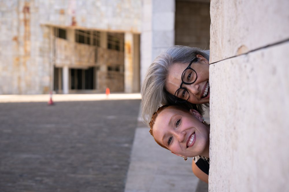 Two women laughing and peeking from behind a stone wall in an urban outdoor setting, businesswomen, Spain.