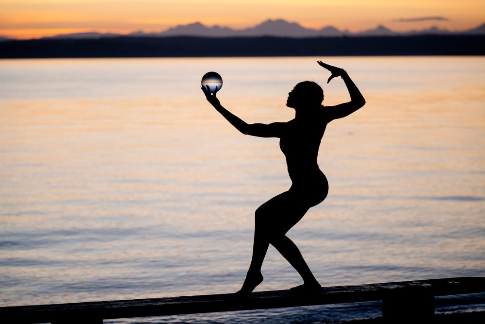 Silhouette of a woman balancing on a beam while holding a glass sphere during sunset by a calm lake, yoga, acrobatics, USA.