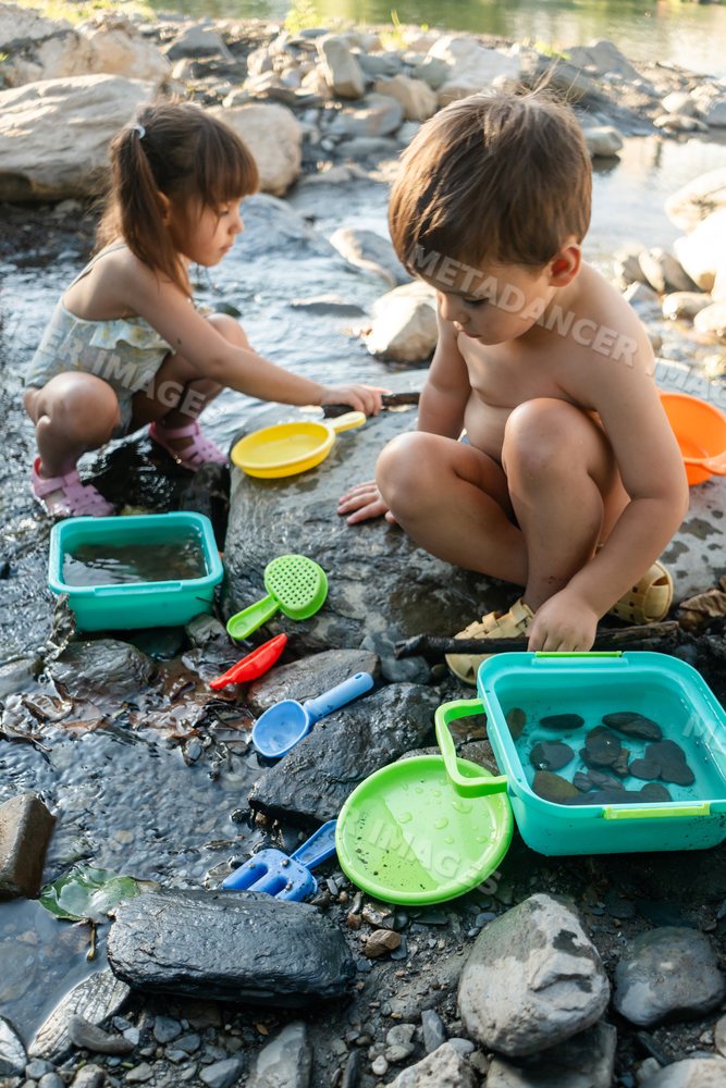 A white boy and girl play with water and toys by a rocky stream