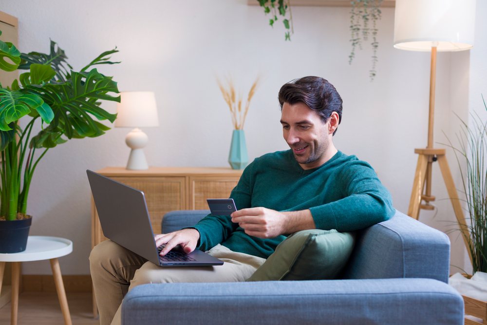 Man sits on a sofa in a modern living room using a laptop while holding a credit card, technology, young adult, home.