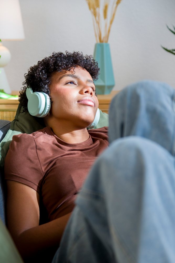 Young woman with curly hair in a brown shirt and blue jeans listens to music with headphones while sitting indoors on a couch at home.