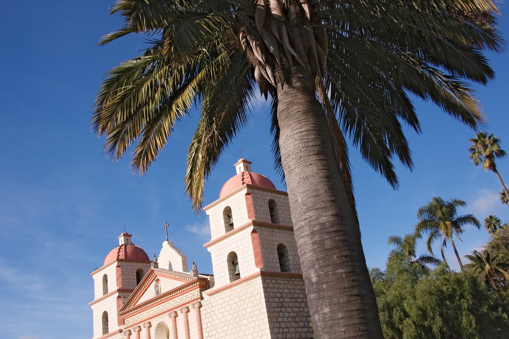 Santa Barbara Mission church with two bell towers and palm trees under a clear blue sky, California, USA.