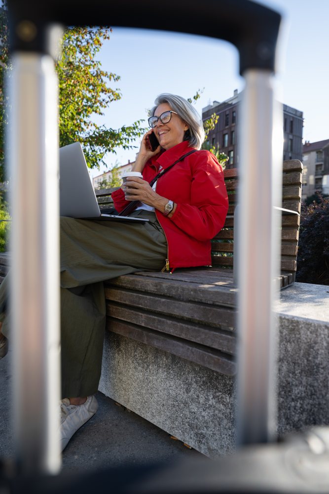 Woman sitting on bench using laptop and phone outdoors in urban park, Spain.
