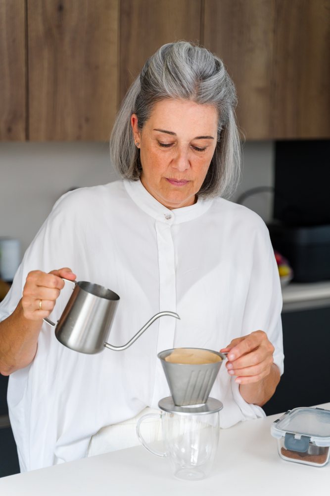 Woman pouring water into a coffee dripper with a paper filter over a glass cup on a kitchen counter, Spain.