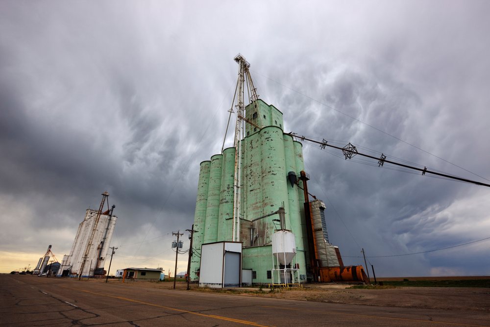 Grain elevators with multiple large silos and machinery under storm clouds beside a rural road, Texas, USA.