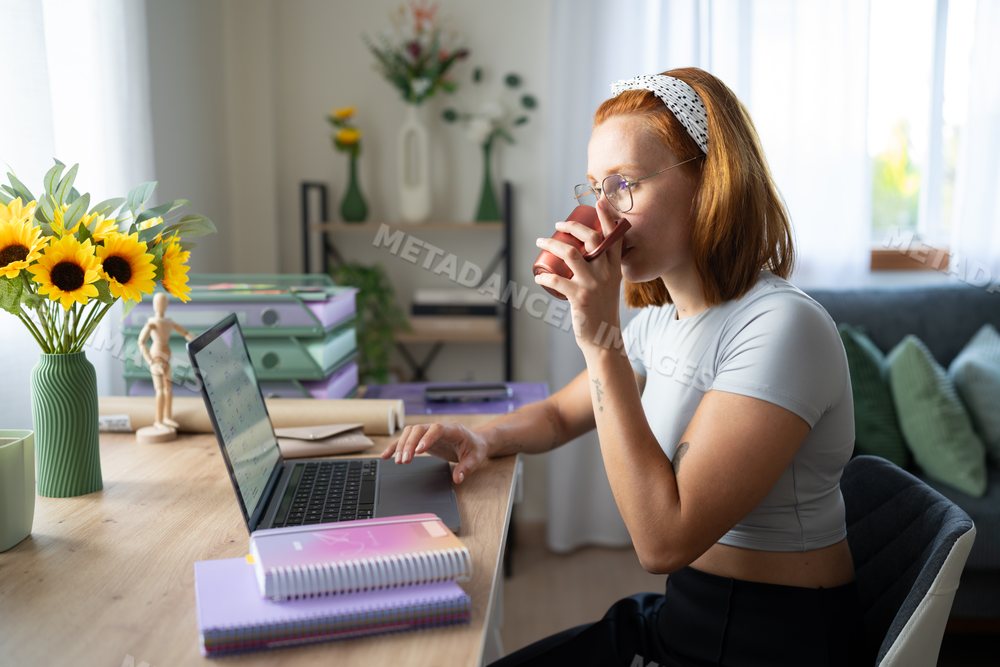A white woman drinks coffee while working on her laptop at home