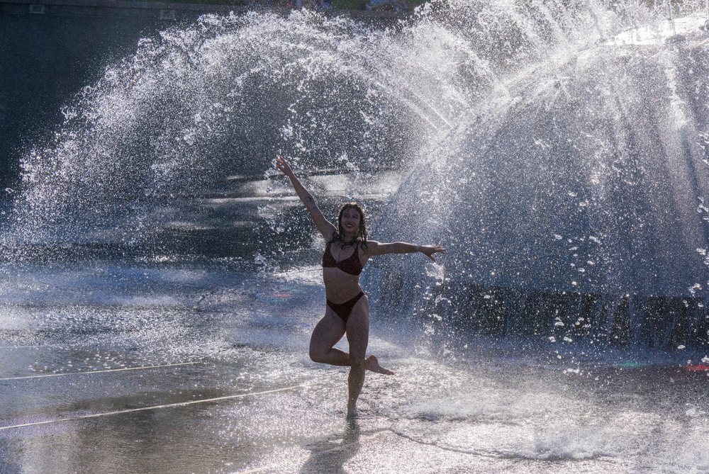 Woman dancing and playing in a water fountain with sprays and splashes at an outdoor public space, partner, acrobatics, yoga, USA.