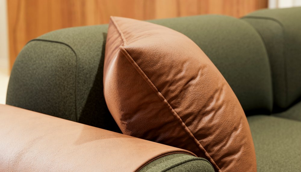 Brown leather pillow resting on the armrest of a green fabric sofa indoors, emphasizing soft textures and cozy seating.