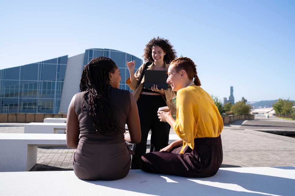 Three women engaging in lively conversation outdoors in an urban park near modern architecture on a sunny day, Spain.