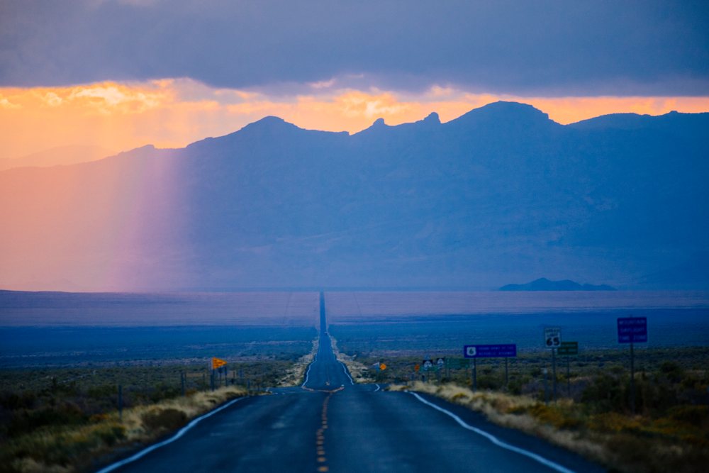 Highway stretches through the Nevada desert with mountains and sun rays at sunset, USA.