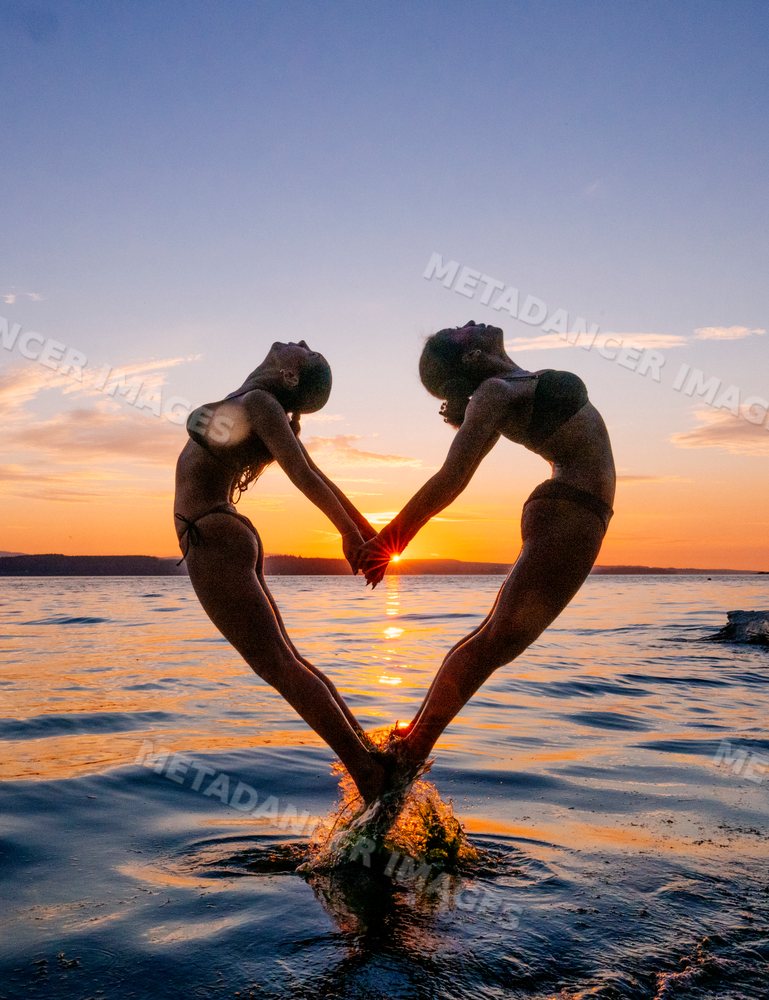Two women perform partner yoga acrobatics pose at sunset over water