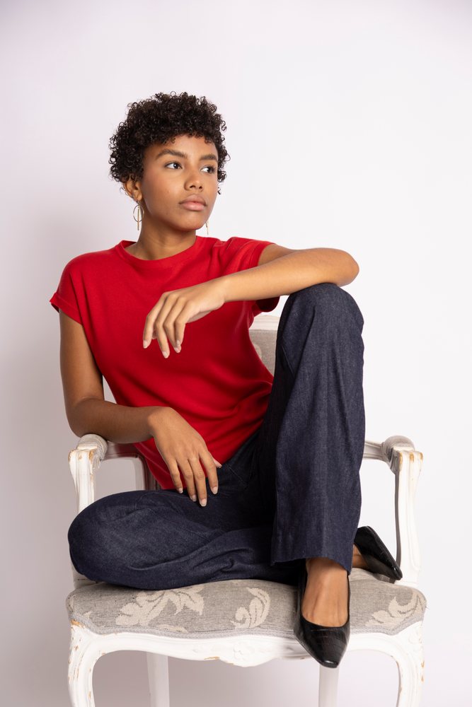 Woman with curly hair sits in an armchair wearing a red shirt and black pants against a white background, fashion, young culture.