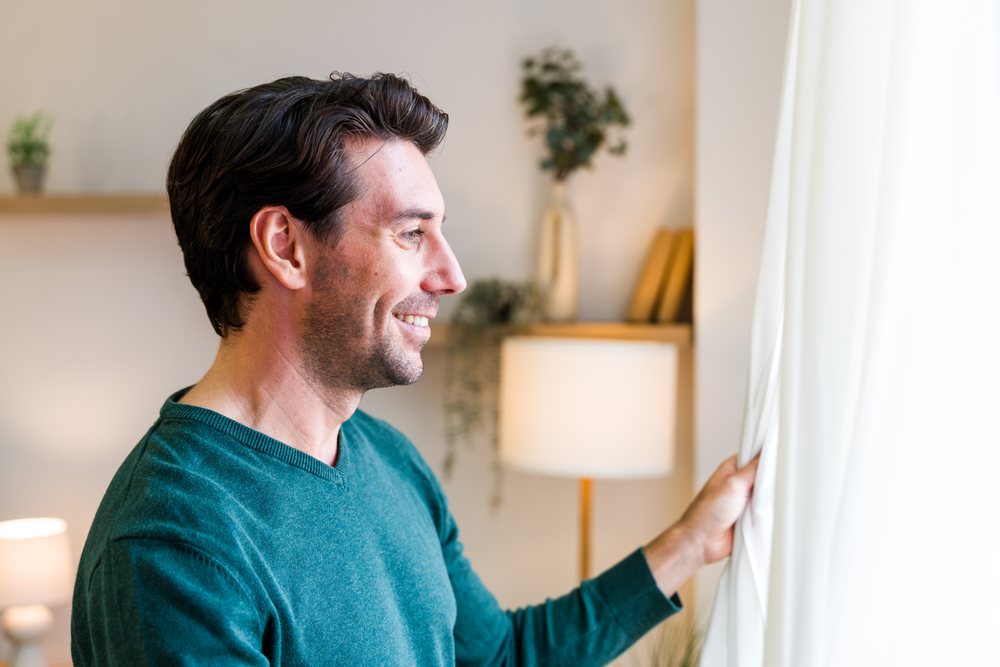 Man smiling as he pulls back a white curtain inside a cozy, well-lit living room interior, Spain.