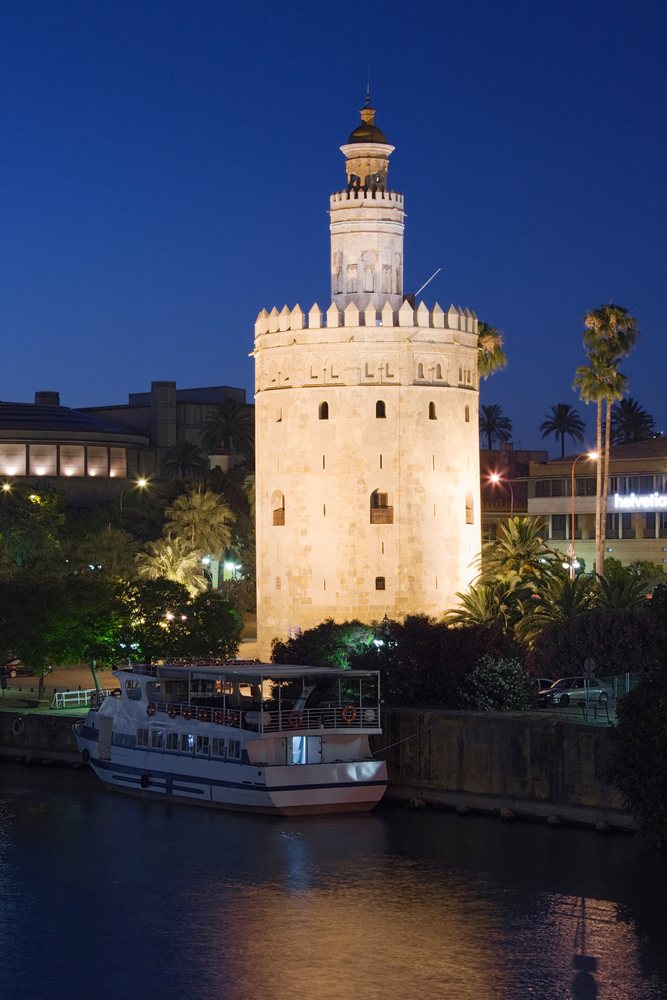 Illuminated Torre del Oro at night along the Guadalquivir River with a moored passenger boat and palm trees, Andalucia, Spain.