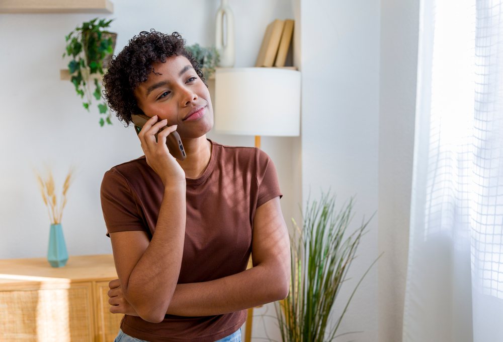 Young woman with curly hair standing indoors talking on a mobile phone near a window with natural light in a modern home.