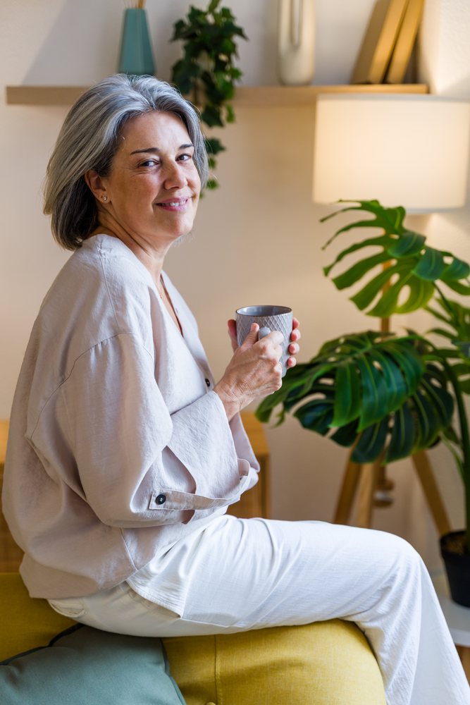 Middle-aged woman with gray hair smiles while holding a mug sitting on a yellow couch in a modern living room, Spain.