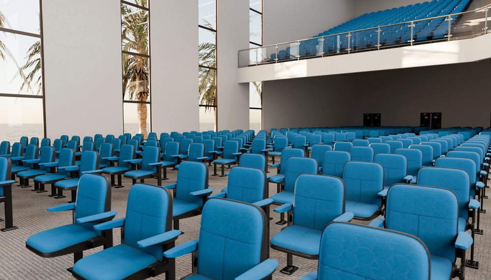 Empty auditorium with rows of blue upholstered chairs, large floor-to-ceiling windows showing palm trees outside, and balcony seating.