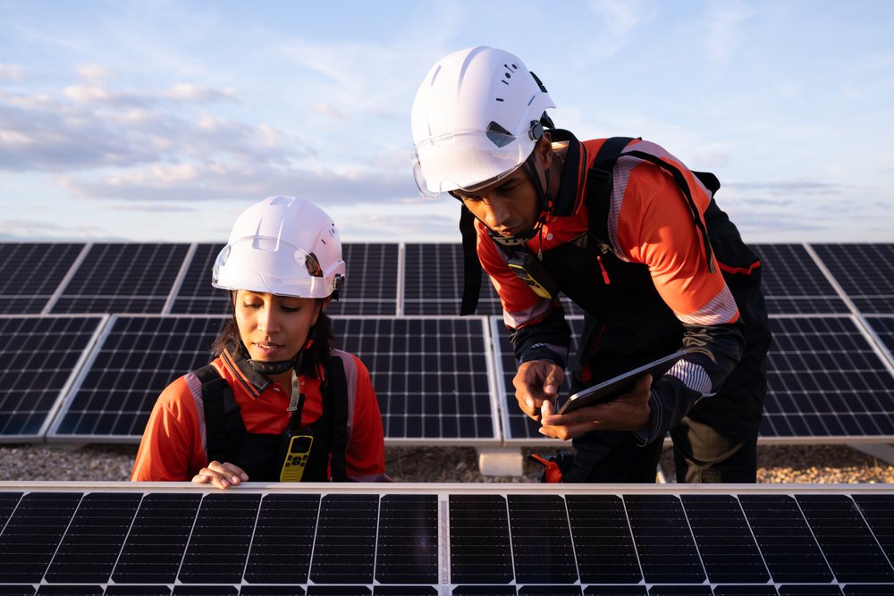 Two workers wearing safety helmets and workwear inspect solar panels outdoors during clear daylight, Madrid, Spain.