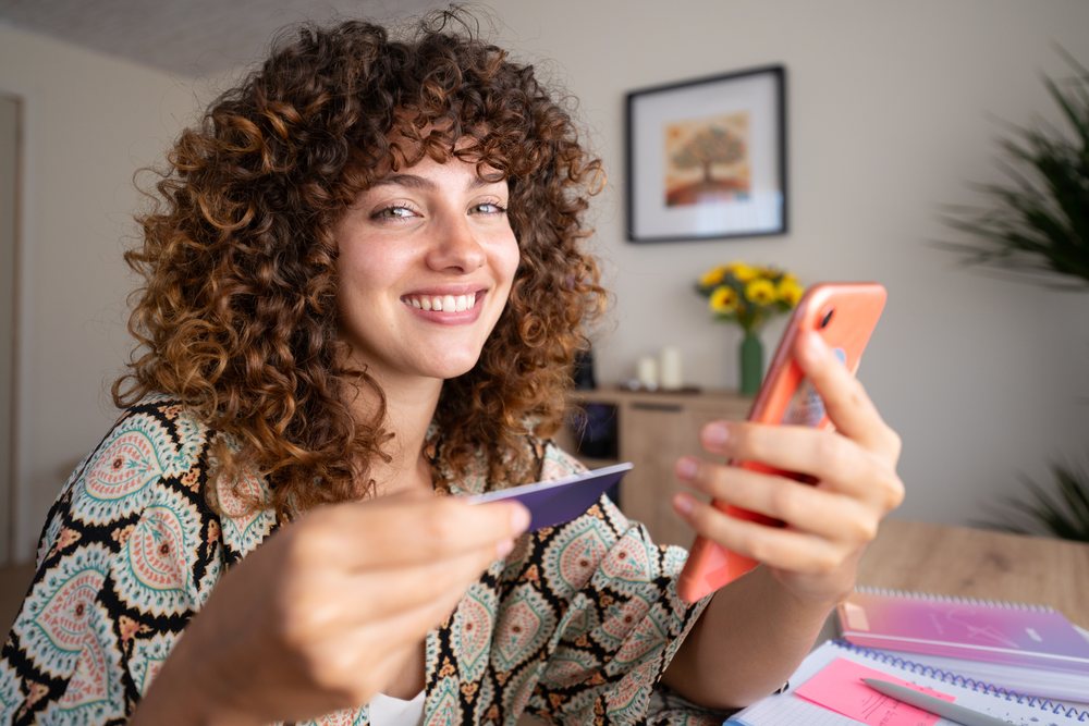 Girl with curly hair smiling, holding a phone and a card inside a home with a desk, notebooks, pen, and flowers in a vase, Spain.