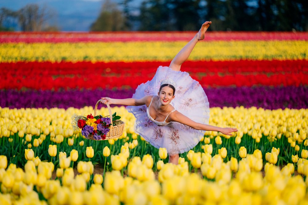 Ballet dancer wearing a tutu and tiara holds a basket of assorted flowers while posing outdoors in a colorful tulip field, USA.