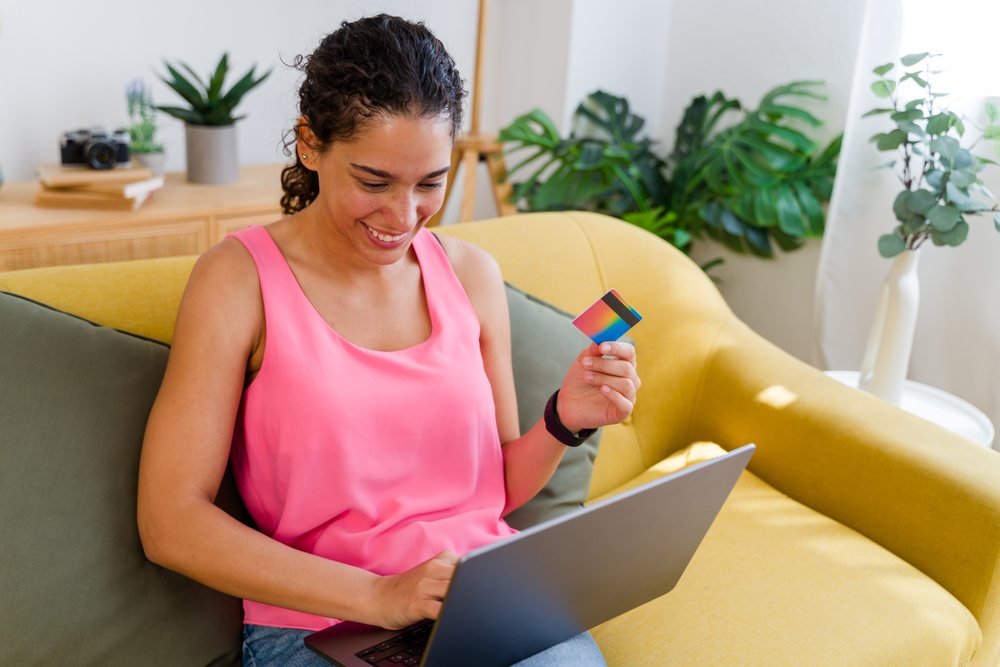 A woman sitting on a yellow couch holds a credit card and uses a laptop to shop online in a bright living room decorated with plants, Spain.