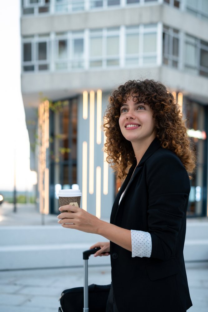 Young business woman holding a coffee cup and pulling a suitcase outdoors by a modern building, Madrid.