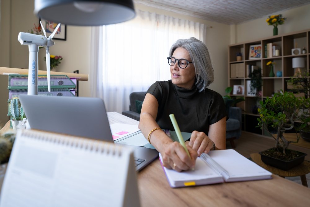Woman writing notes in a notebook at her home office desk with a laptop, wind turbine model, cactus plant, and files nearby, Spain.