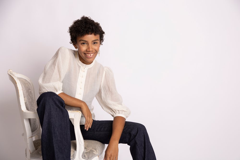 Young woman with curly hair smiles sitting on a vintage armchair against a white studio background wearing a white blouse and dark blue pants, fashion, young culture.