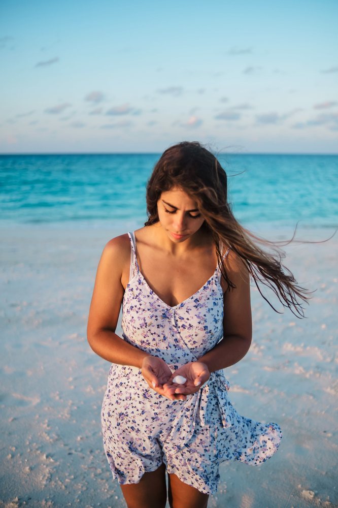 A woman in a floral summer dress stands on a beach holding a seashell with the ocean in the background in the, Maldives.