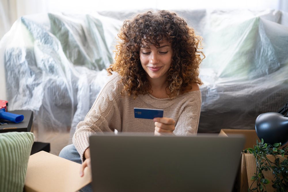 Woman with curly hair sits on a plastic-covered sofa holding a credit card and using a laptop amid moving boxes indoors, Spain.