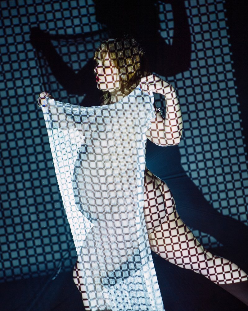 Woman holding semi-transparent white fabric with checkered light pattern projecting shadows and silhouette in an artistic indoor setting, pregnancy.
