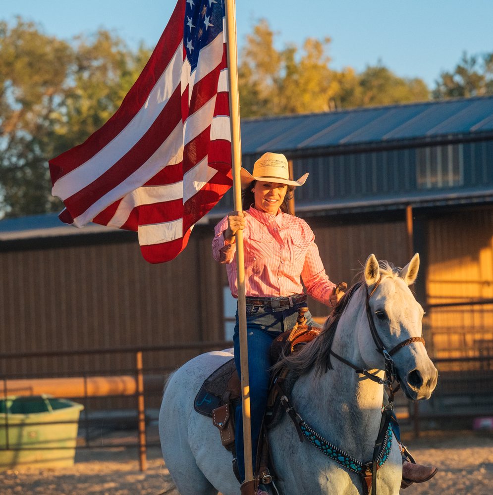 Woman wearing a cowboy hat and striped shirt rides a white horse outdoors near a metal stable in the Southwest USA while holding an American flag on a flagpole.
