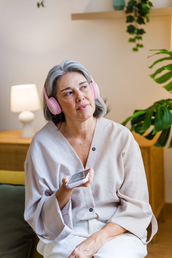 Senior woman with gray hair wears pink headphones and holds a phone indoors in a living room with houseplant and lamp, Spain.