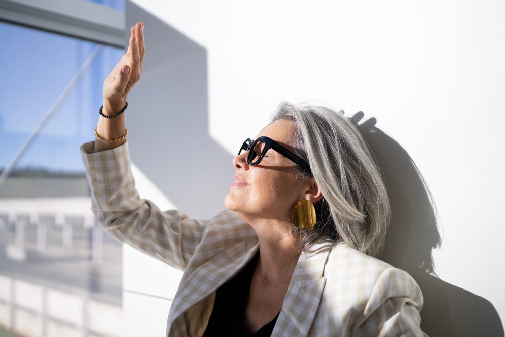 Mature woman wearing black sunglasses and a checkered beige blazer stands outdoors in bright sunlight casting a shadow on a white wall, businesswoman, traveler, Spain, Madrid.