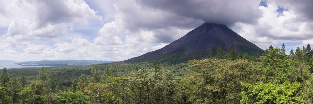 Arenal Volcano, a conical volcanic mountain, rises above dense tropical forest under a partly cloudy sky.