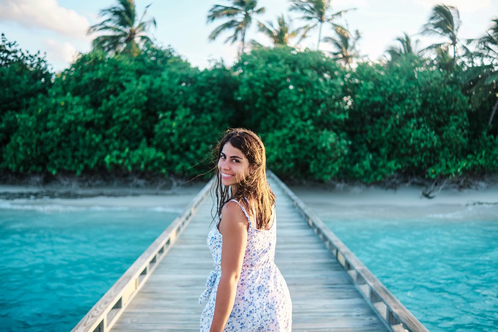 A young woman in a blue floral dress smiles as she walks along a wooden pier leading to a tropical beach in the, Maldives.